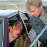 Scena z filmu o zabijaniu: ranny mężczyzna z krwią na twarzy siedzi w samochodzie, obok niego stoi drugi mężczyzna z bronią.