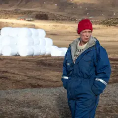 Kobieta w niebieskim kombinezonie i czerwonej czapce stoi na polu, daleko od Reykjaviku, obok sterty białych bel siana.