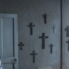 Przerażona kobieta w niebieskim płaszczu opiera się o ścianę pokrytą wzorzystą tapetą, na której wisi wiele krzyży. Czy to znak obecności duchów z Enfield?