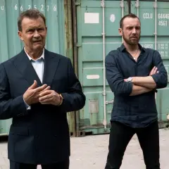 Dwóch mężczyzn w garniturach, jeden z nich z rękami złożonymi na piersi, jakby zastanawiał się, jak zostać gangsterem. Obsada serialu.