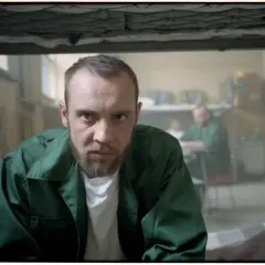 Mężczyzna w zielonej kurtce, jak z filmu "Jak zostałem gangsterem. Historia prawdziwa", patrzy wprost. W tle inni więźniowie.