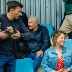 Grupa ludzi na trybunach, wśród nich starszy pan w tweedowej marynarce, wspierany przez młodszego mężczyznę. Scena jak z najlepszych polskich filmów.