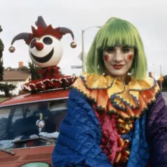 Kobieta w zielonej peruce i stroju klauna z serduszkami na policzkach, obok czerwonego samochodu z zabawną kukłą na dachu, scena z filmu Roberta Altmana.