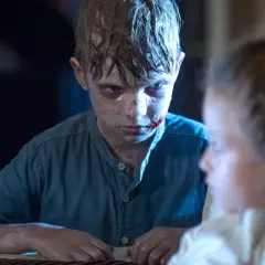 Chłopiec z ranami na twarzy patrzy na coś po tamtej stronie drzwi. Obok siedzi dziecko.