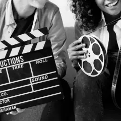 Dwie osoby trzymają klaps filmowy i rolkę taśmy filmowej. Zastanawiasz się, ile zarabia producent filmowy?