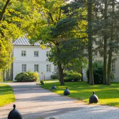 Biały dworek otoczony zielenią, z alejką i ozdobnymi kulami. Miejsce, gdzie nagrywano "Nad Niemnem".