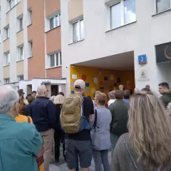 Grupa ludzi stoi przed blokiem mieszkalnym z kolorową ścianą i numerem 2.