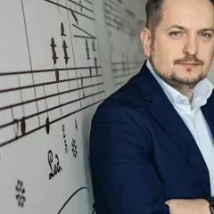 Mężczyzna w granatowej marynarce, oparty o ścianę z nutami. Wygląda jak człowiek do wszystkiego, gotowy na każde wyzwanie.