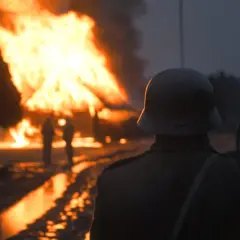 Żołnierz w hełmie obserwuje płonące chaty w świetle dnia. W tle widać innych żołnierzy i druty kolczaste.
