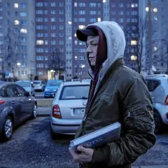 Młody mężczyzna w bluzie z kapturem i kurtce bomberce trzyma w ręku płytę. W tle widać blokowisko i zaparkowane samochody, jakby scena z filmu o innych ludziach.