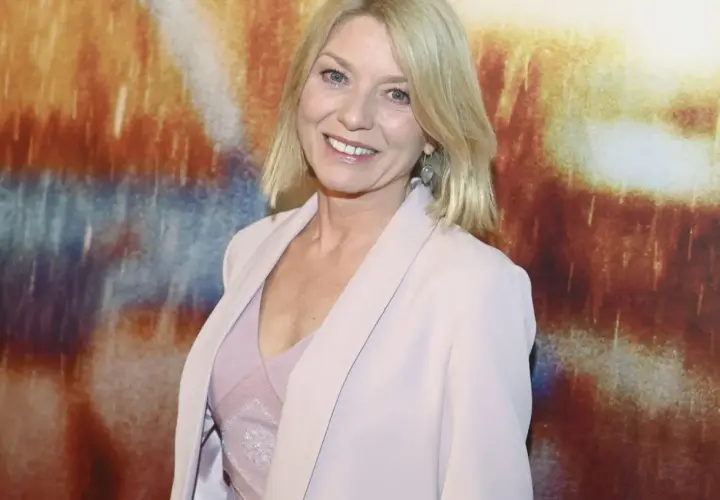 Uśmiechnięta kobieta z blond włosami, w jasnoróżowym żakiecie, na tle abstrakcyjnego obrazu. Może to być kadr z filmu.
