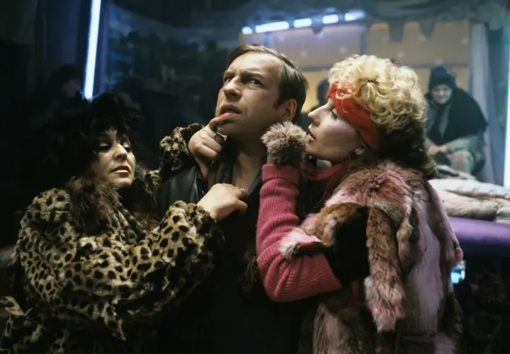 Scena z filmu Piotra Szulkina: mężczyzna w futrze w panterkę i różowym futrze, otoczony przez dwie kobiety w ekstrawaganckich strojach.