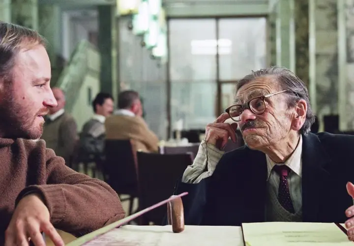 Dwóch mężczyzn rozmawia przy stole. Jeden z nich, starszy, w okularach, gestykuluje. Scena z jednego z filmów Krzysztofa Krauzego.