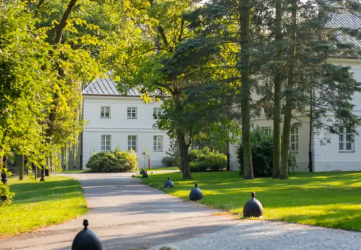 Biały dworek otoczony zielenią, z alejką i ozdobnymi kulami. Miejsce, gdzie nagrywano "Nad Niemnem".