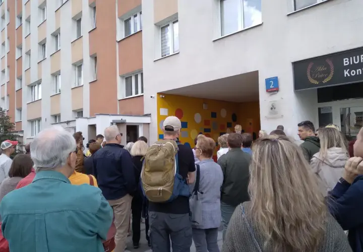 Grupa ludzi stoi przed blokiem mieszkalnym z kolorową ścianą i numerem 2.