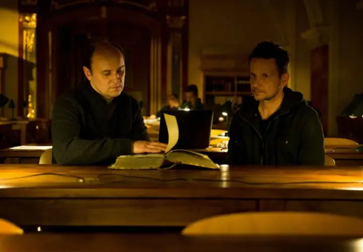 Dwóch mężczyzn przy stole w bibliotece, jeden czyta starą księgę. Scena z filmu "Sługi Boże".