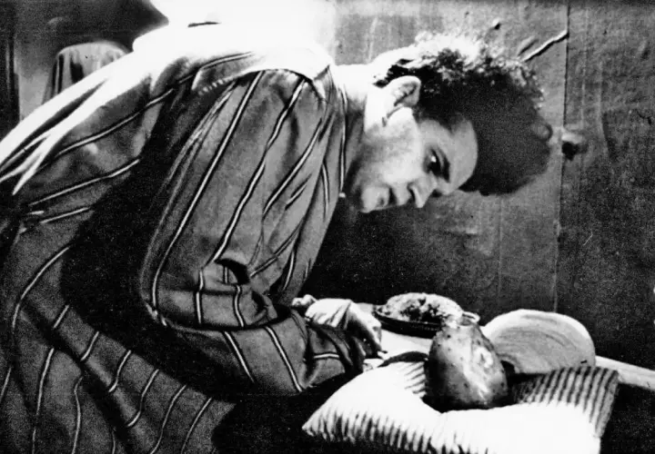 Młody David Lynch w pasiastym szlafroku pochyla się nad stołem, na którym leżą dziwne przedmioty.