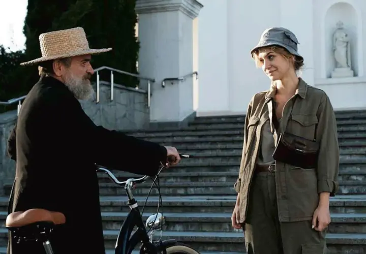 Scena z filmu Jacka Bromskiego: mężczyzna w słomkowym kapeluszu i czarnej sutannie przy rowerze rozmawia z kobietą w wojskowym kapeluszu.