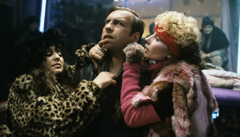 Scena z filmu Piotra Szulkina: mężczyzna w futrze w panterkę i różowym futrze, otoczony przez dwie kobiety w ekstrawaganckich strojach.