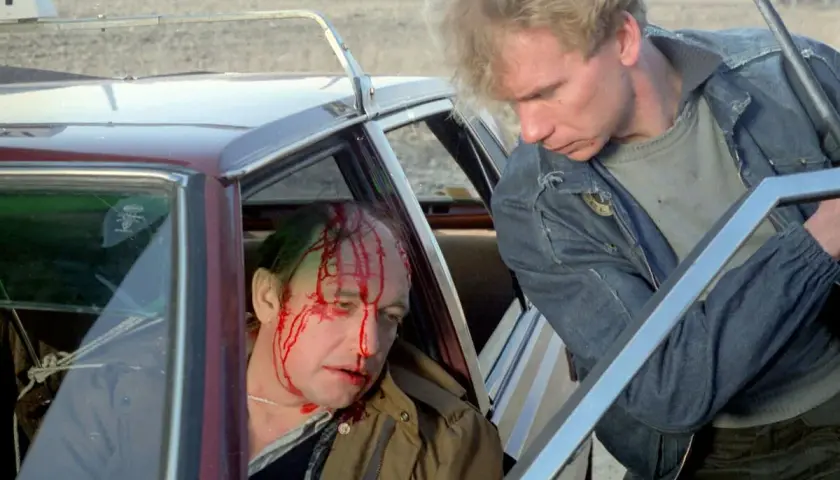 Scena z filmu o zabijaniu: ranny mężczyzna z krwią na twarzy siedzi w samochodzie, obok niego stoi drugi mężczyzna z bronią.