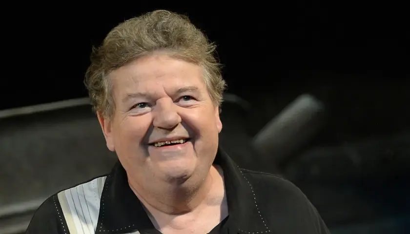 Uśmiechnięty Robbie Coltrane, który grał Hagrida, w czarnej koszuli z białymi pasami.