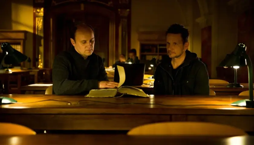 Dwóch mężczyzn przy stole w bibliotece, jeden czyta starą księgę. Scena z filmu "Sługi Boże".