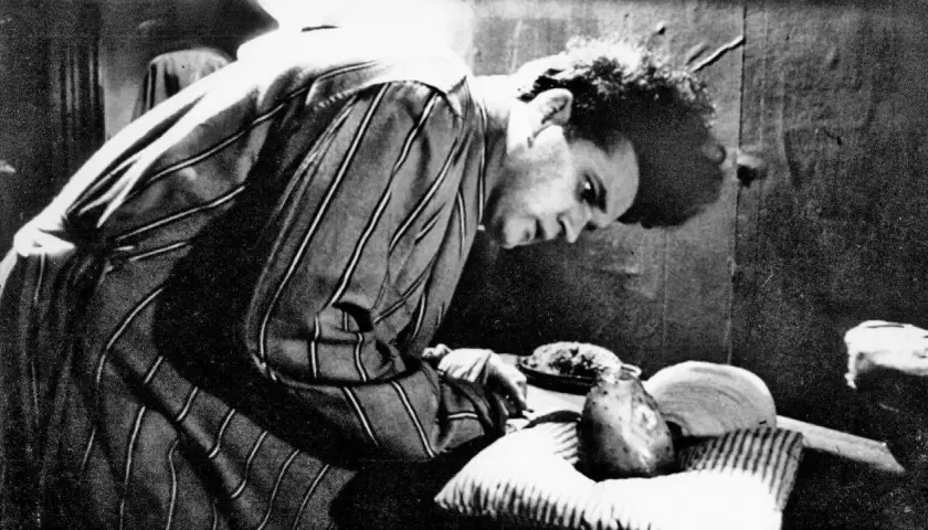 Młody David Lynch w pasiastym szlafroku pochyla się nad stołem, na którym leżą dziwne przedmioty.