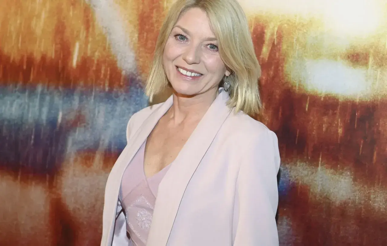 Uśmiechnięta kobieta z blond włosami, w jasnoróżowym żakiecie, na tle abstrakcyjnego obrazu. Może to być kadr z filmu.