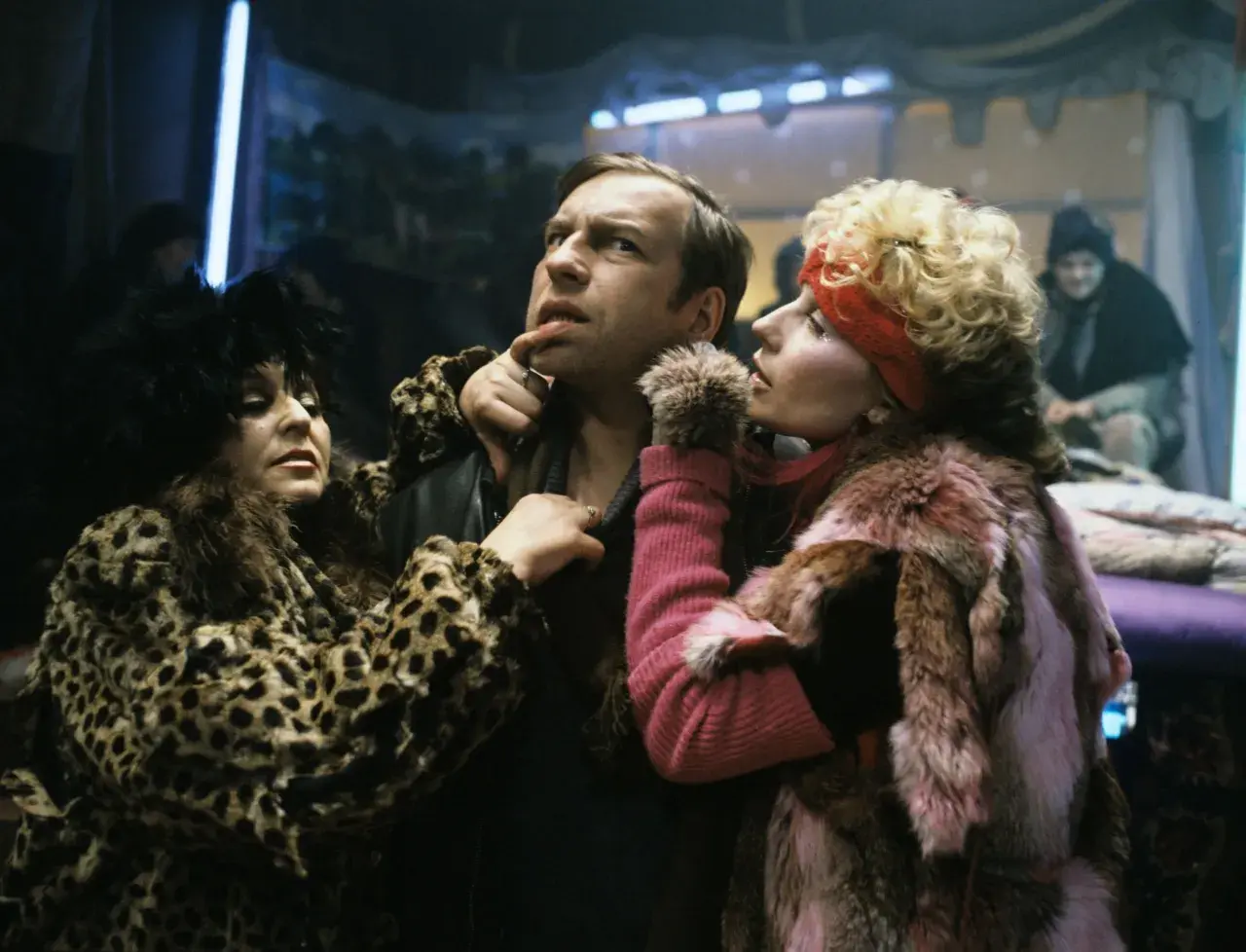 Scena z filmu Piotra Szulkina: mężczyzna w futrze w panterkę i różowym futrze, otoczony przez dwie kobiety w ekstrawaganckich strojach.