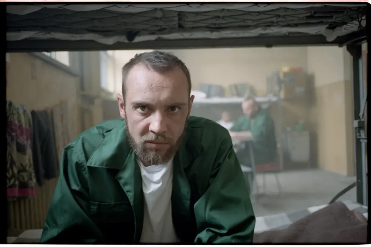 Mężczyzna w zielonej kurtce, jak z filmu "Jak zostałem gangsterem. Historia prawdziwa", patrzy wprost. W tle inni więźniowie.