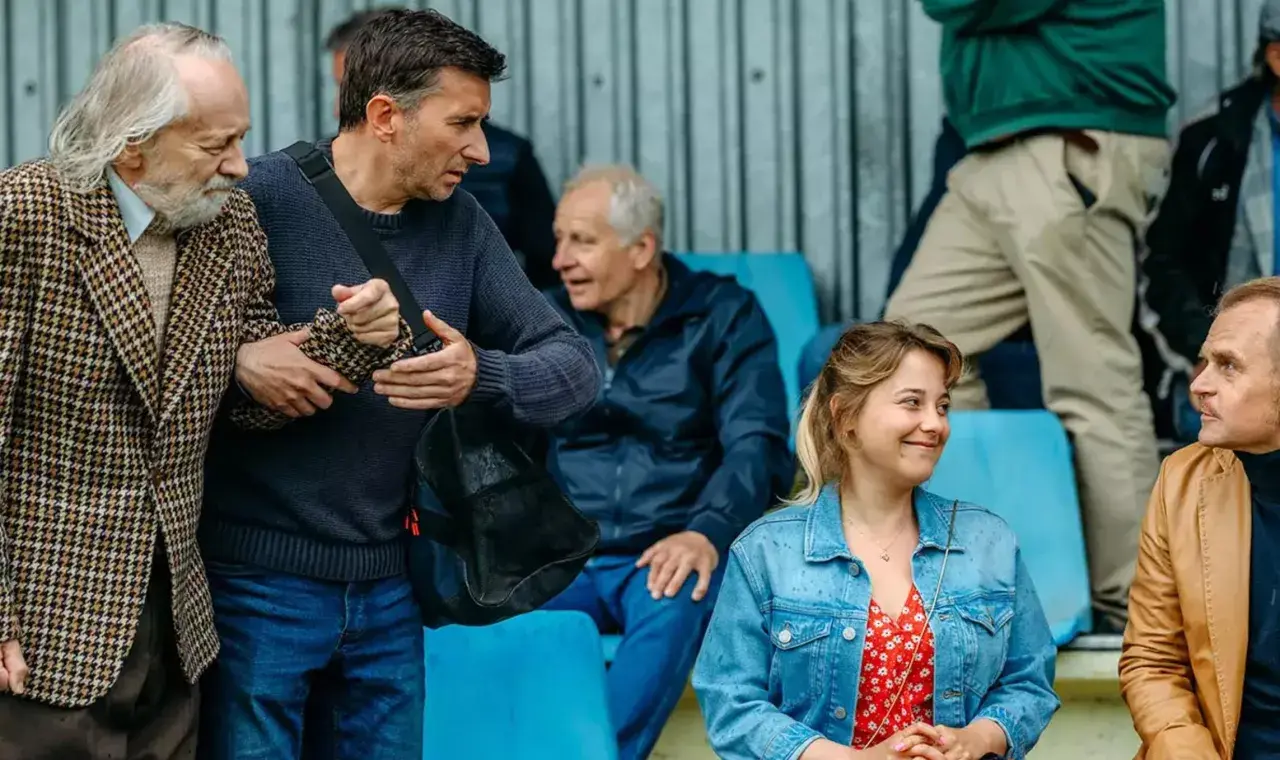Grupa ludzi na trybunach, wśród nich starszy pan w tweedowej marynarce, wspierany przez młodszego mężczyznę. Scena jak z najlepszych polskich filmów.
