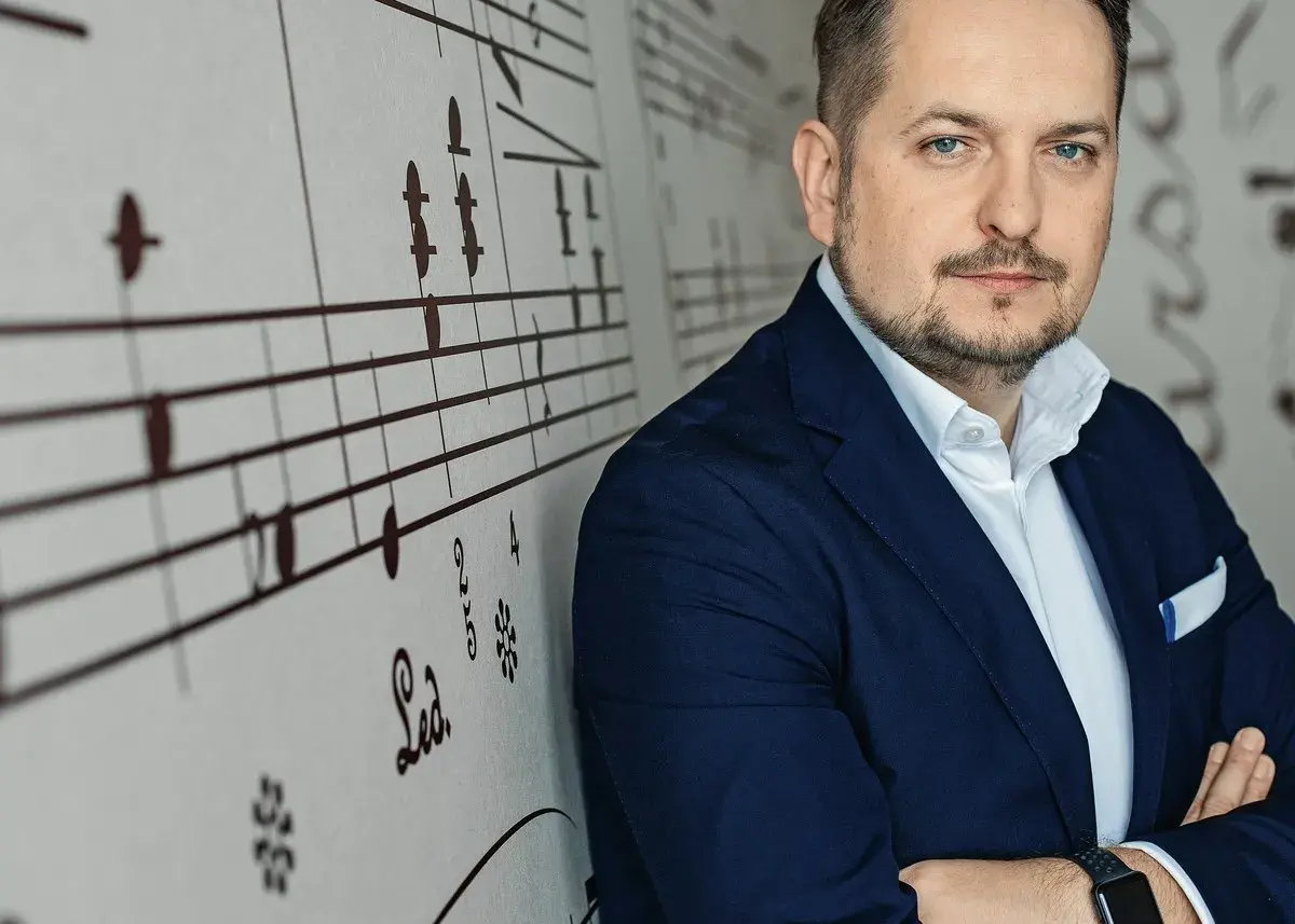 Mężczyzna w granatowej marynarce, oparty o ścianę z nutami. Wygląda jak człowiek do wszystkiego, gotowy na każde wyzwanie.