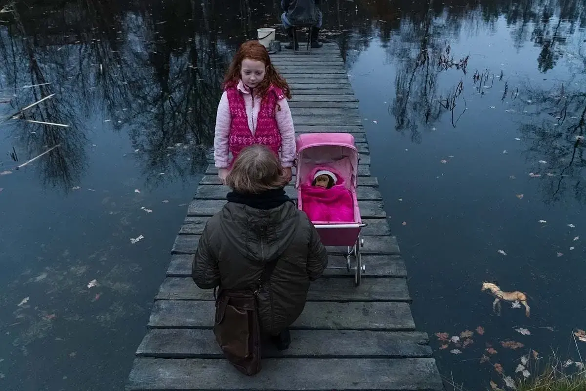 Dwie dziewczynki na drewnianym pomoście, jedna z wózkiem dla lalek, druga obok. Scena jak z filmu "ciemno prawie noc".
