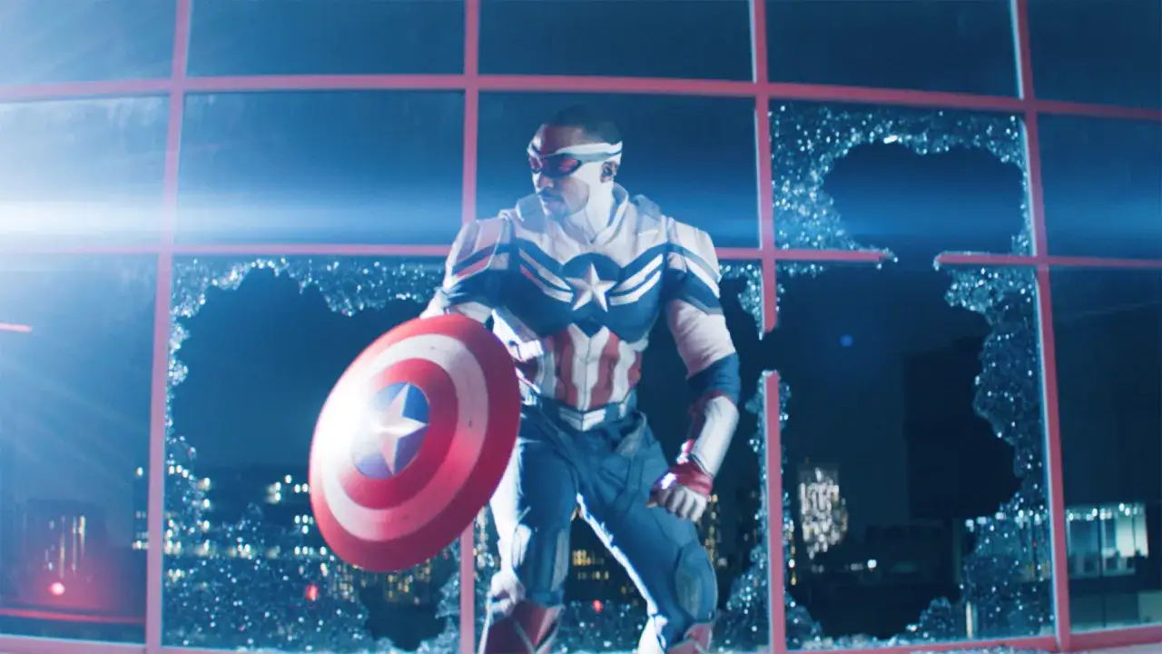 Nowy Kapitan Ameryka filmu, Sam Wilson, w swoim stroju, z tarczą, stoi przed rozbitym oknem.