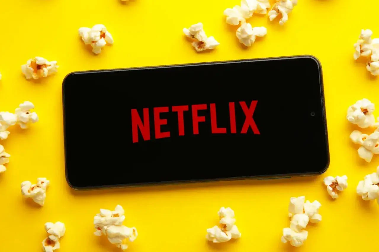 Telefon z logo Netflix otoczony popcornem na żółtym tle. Idealny zestaw na wieczór z filmem warty obejrzenia.