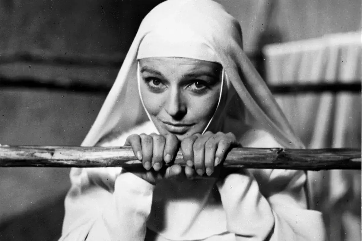Lucyna Winnicka w filmach, jako zakonnica, opiera się o drewniany drążek, patrząc wprost na widza.