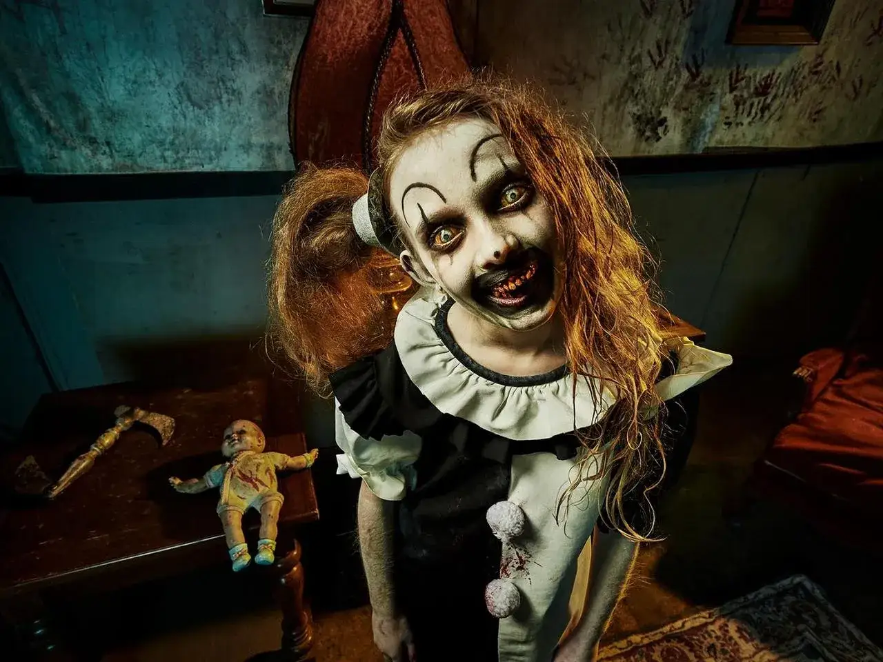 Przerażający klaun z horroru, jak z Terrifier 3, z makabrycznym uśmiechem i żółtymi oczami, stoi obok lalek i siekiery.