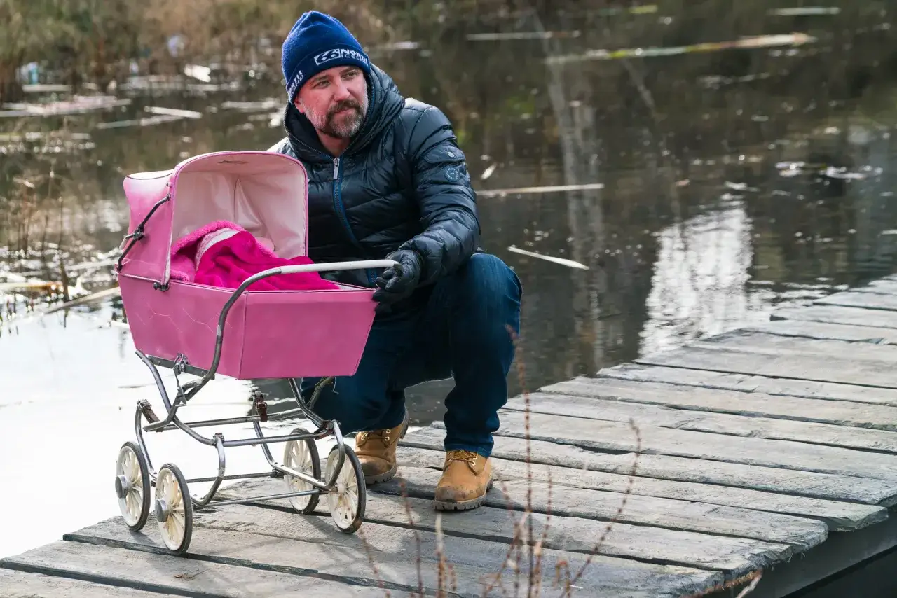 Mężczyzna w czapce i kurtce kuca przy różowym wózku na drewnianym pomoście. Scena jak z filmu 