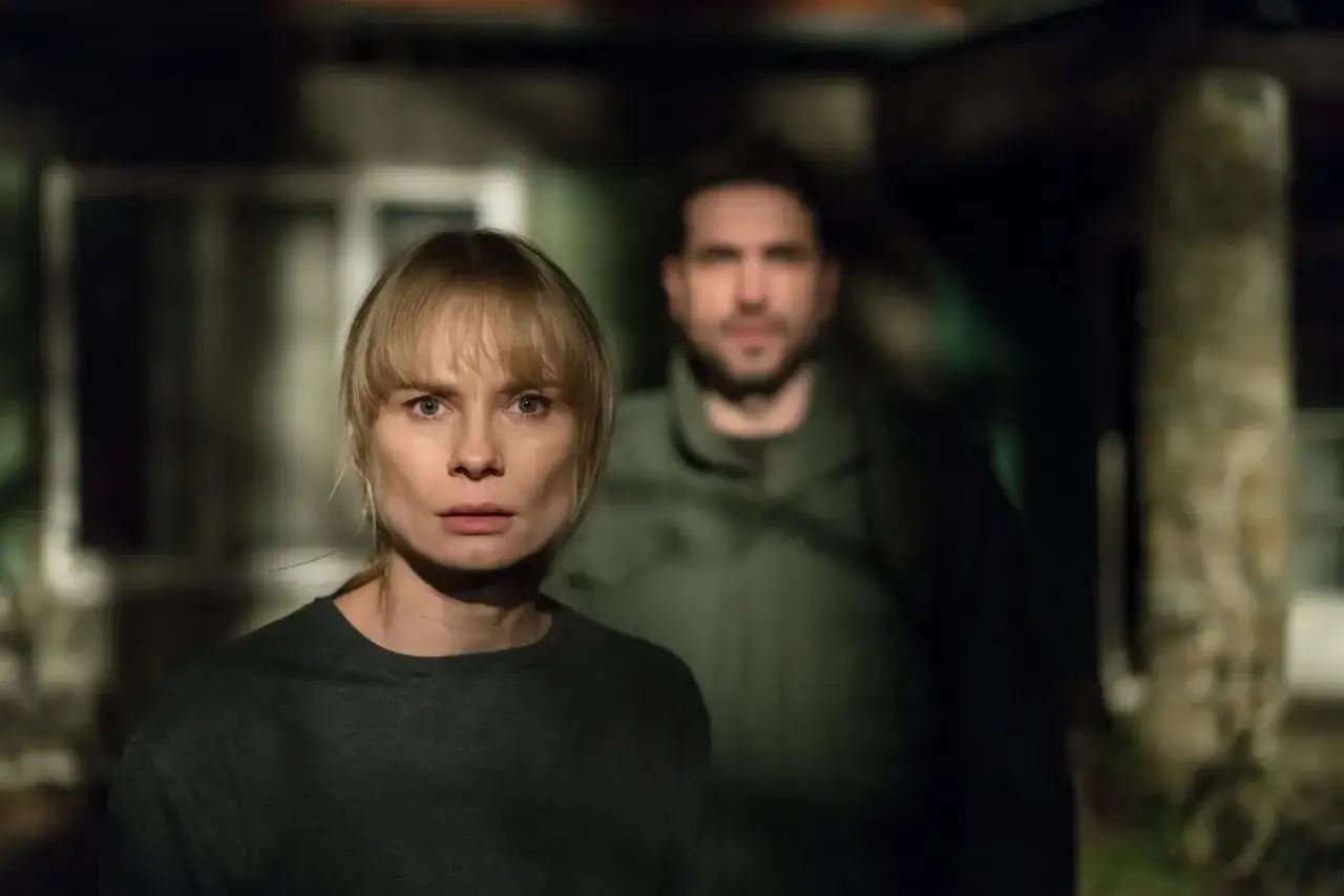 Kobieta z przerażeniem patrzy w obiektyw, za nią w mroku stoi mężczyzna. Scena z filmu 