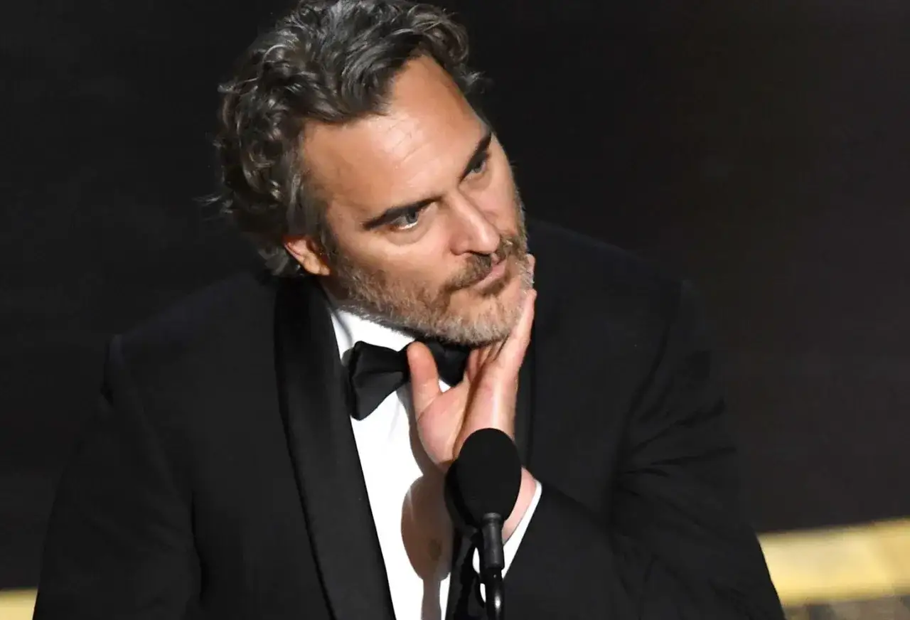 Joaquin Phoenix, który grał Jokera w filmie Joker, w czarnym smokingu z muszką, dotyka swojej brody.